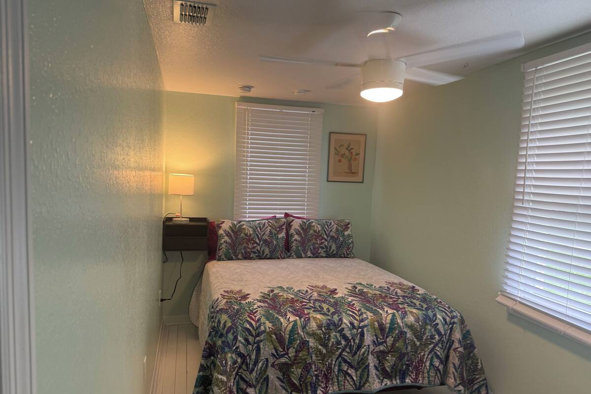 A bedroom with a bed, a nightstand, a lamp, a ceiling fan, and a window with blinds.