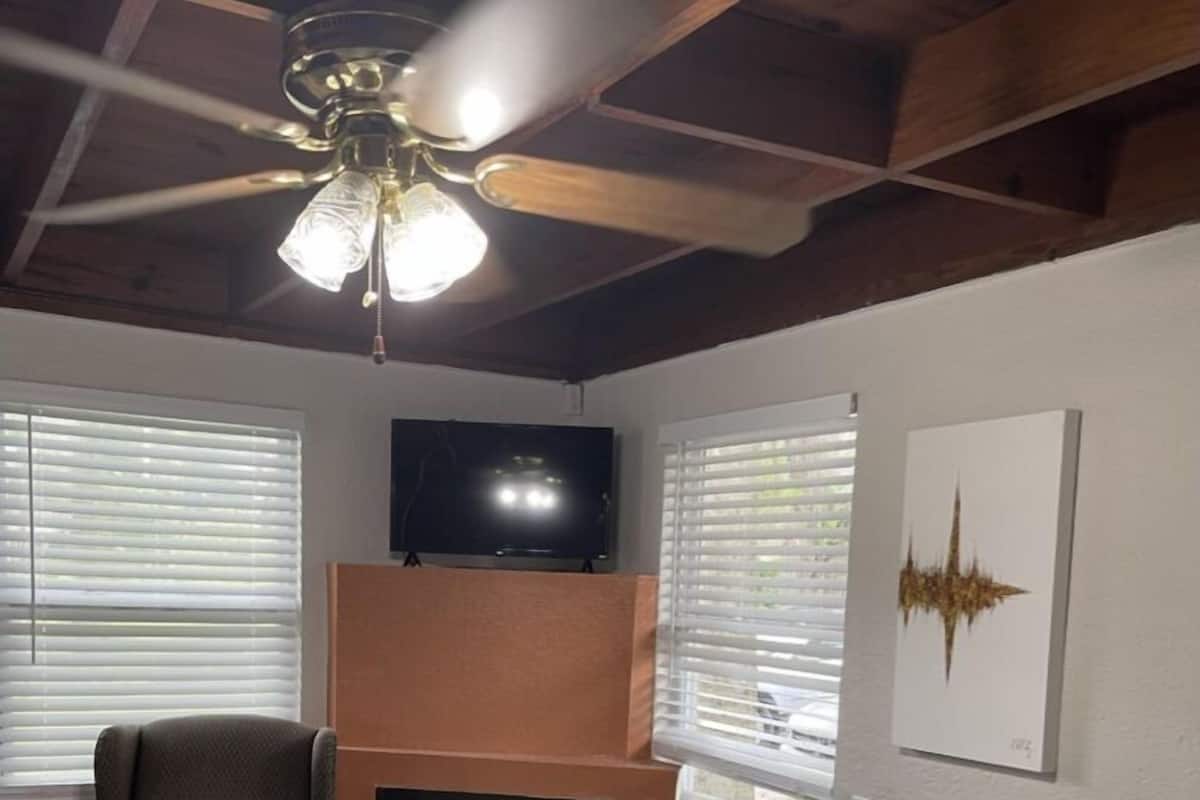 A bedroom with a bed, a chair, a fireplace, a ceiling fan, and a TV on the wall.