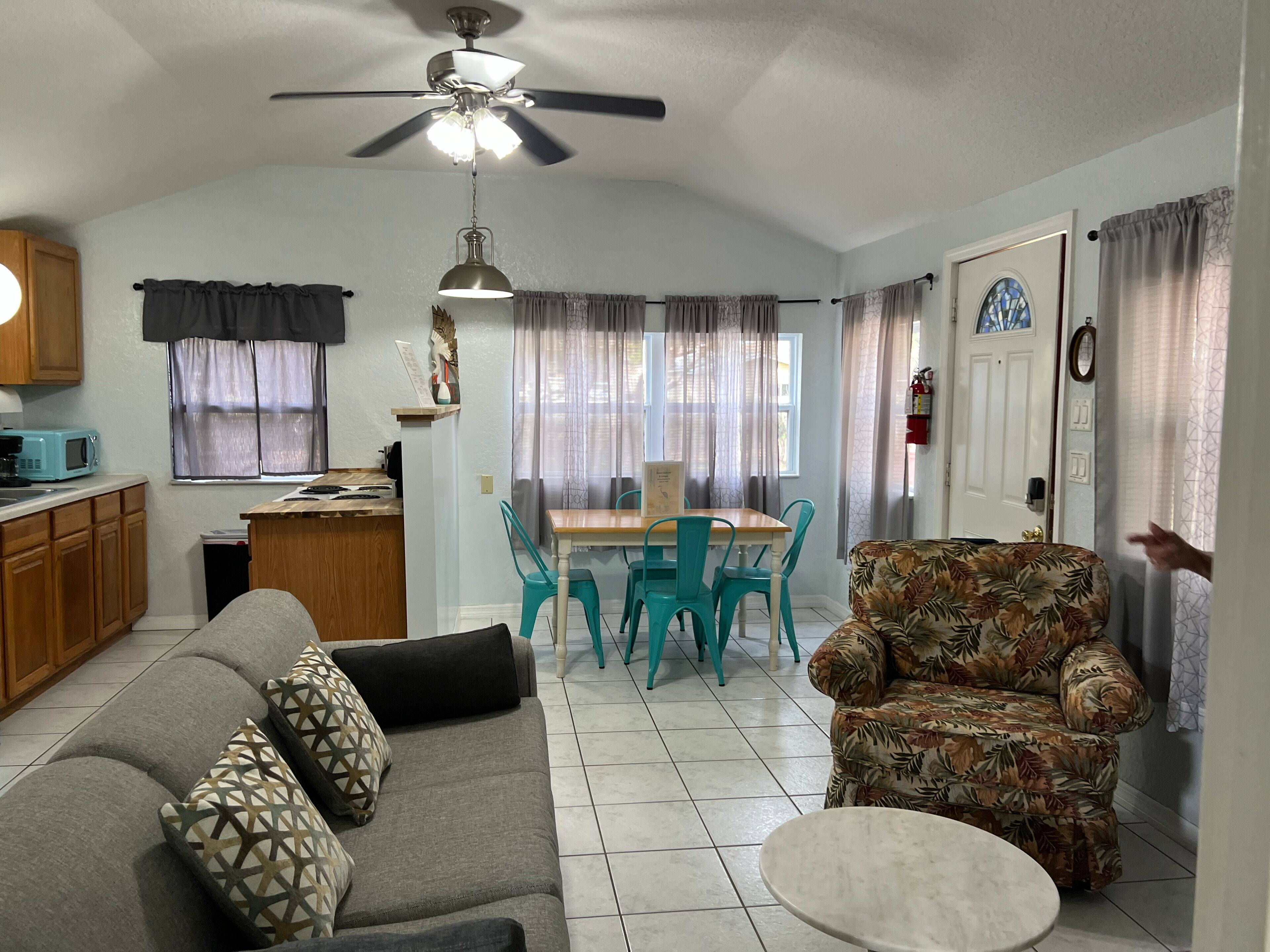 A living room with a sofa, a patterned armchair, a dining table with chairs, a ceiling fan, and a kitchen area with cabinets and a microwave.