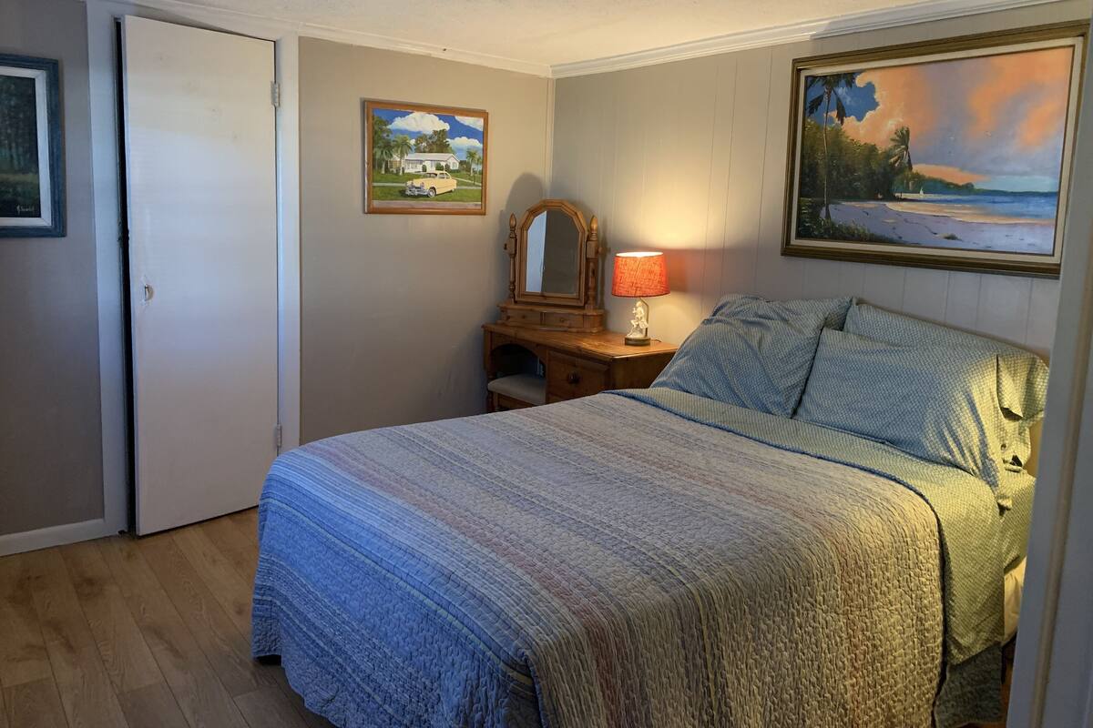 A bedroom with a bed, a nightstand with a lamp, a dresser, a mirror, and two framed pictures on the wall.