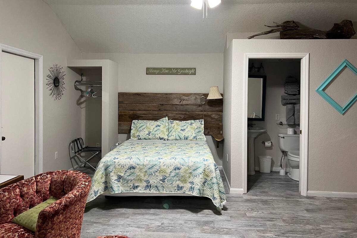 A bedroom with a bed, a ceiling fan, a bathroom visible through an open door, and a decorative sign on the wall.
