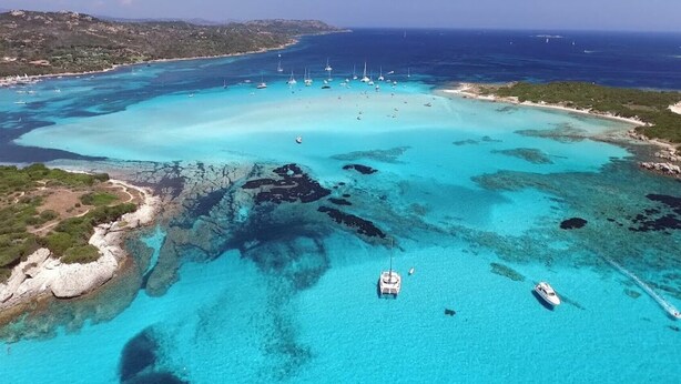 An aerial view of a coastal area with clear turquoise waters, boats, and a rocky shoreline. - Yacht "BAVARIA 37 SPORT "RIBELLE"" in Sardinia - with a sporty design (Olbia)