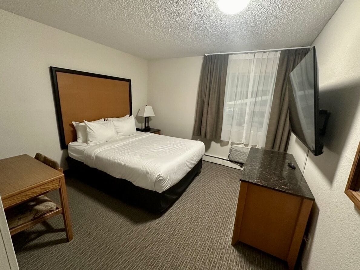 A hotel room with a bed, a desk, a chair, a lamp, a TV, and a window with curtains.