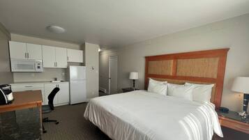 A hotel room with a bed, a desk, a chair, a microwave, a refrigerator, and a nightstand with a lamp.