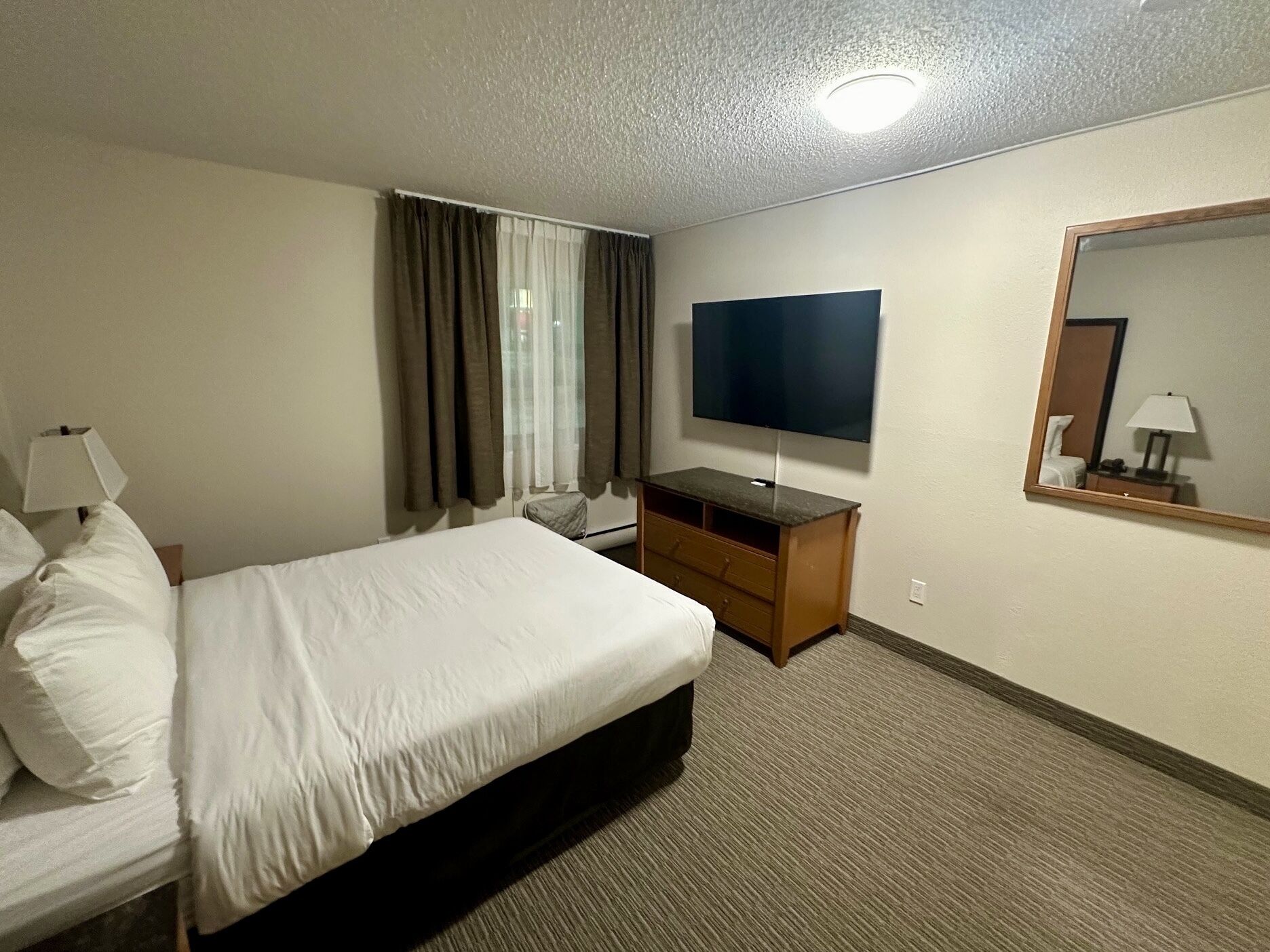A hotel room with a bed, a TV, a dresser, a mirror, and a window with curtains.
