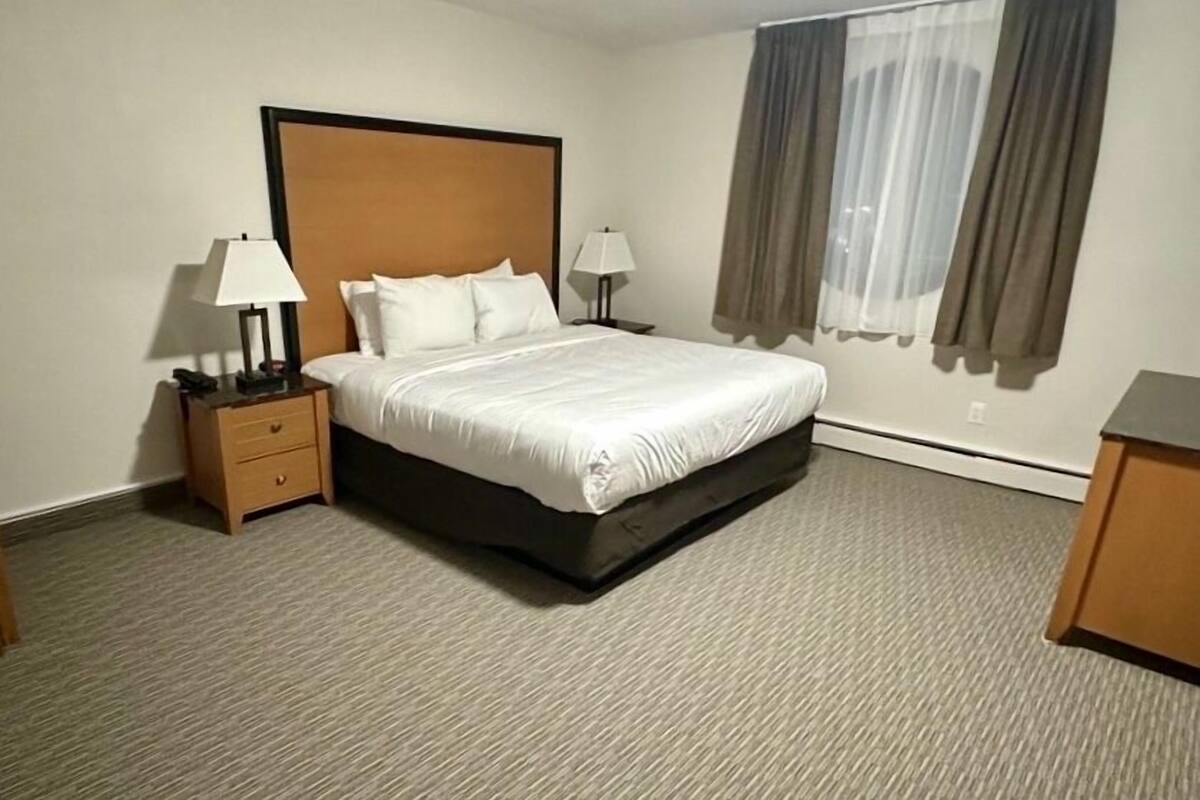 A hotel room with a large bed, two bedside tables with lamps, a window with curtains, and a carpeted floor.