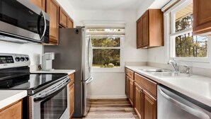 A modern kitchen with wooden cabinets, stainless steel appliances, a window with a view of trees, and a white countertop. - Sound Sanctuary: Canal Front w/ Dock, Dog-Friendly (Southern Shores)