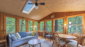 Un intérieur de chalet chaleureux avec un canapé, une table à manger et des chaises, de grandes fenêtres donnant sur la forêt et une lucarne.