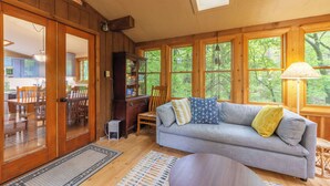 L’intérieur chaleureux d’un chalet avec un coin repas en bois, un canapé agrémenté de coussins et de grandes fenêtres offrant une vue sur la forêt.