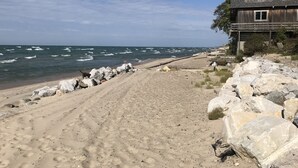 Une plage bordée d’un rivage rocheux, une maison en bois avec un toit à pignon et une jetée qui s’avance dans l’océan.