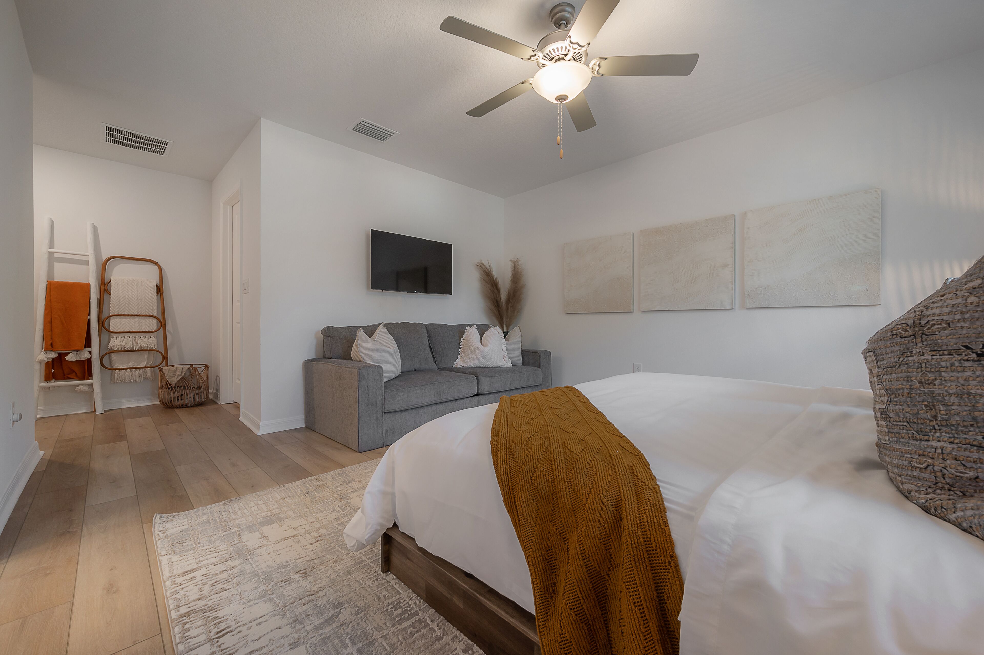 A bedroom with a bed, a grey sofa, a ceiling fan, and a flat-screen TV.