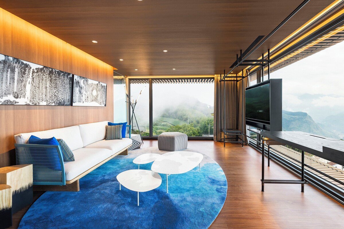 A modern living room with a sofa, ottoman, and coffee table, a large TV, and a view of mountains.