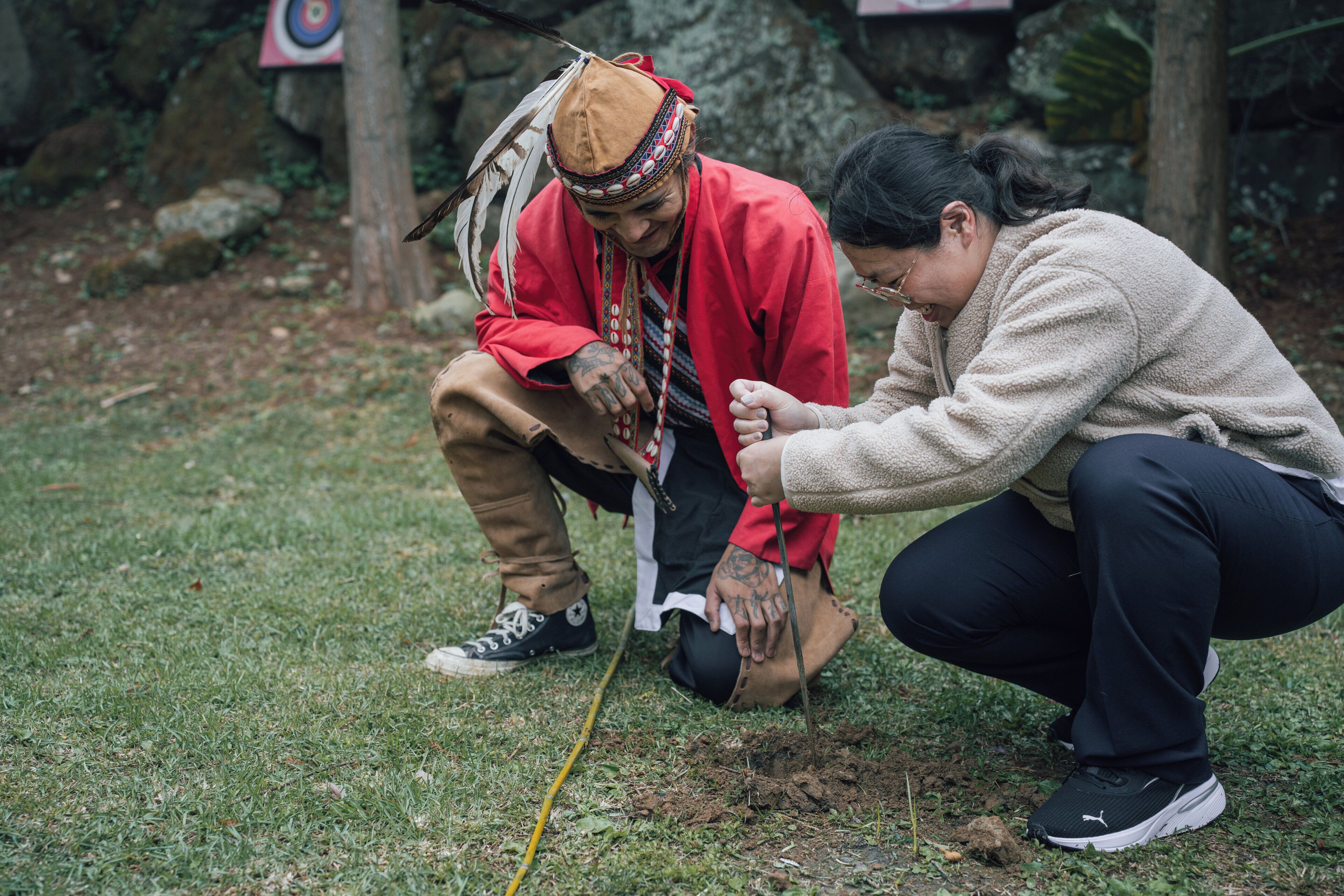 Two individuals in an outdoor setting with grass and rocks, one wearing a red jacket and a hat, and the other in a beige sweater.