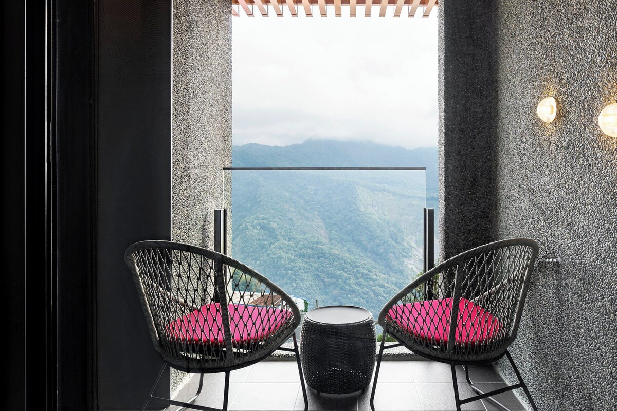 A balcony with two wicker chairs and a small table, overlooking a mountainous landscape.
