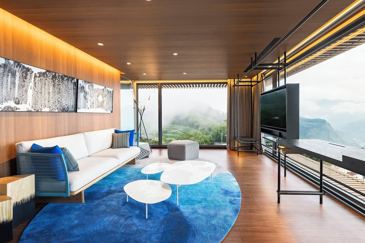 Ein modernes Wohnzimmer mit Sofa, Hocker und Couchtisch, einem großen Fernseher und Blick auf Berge.