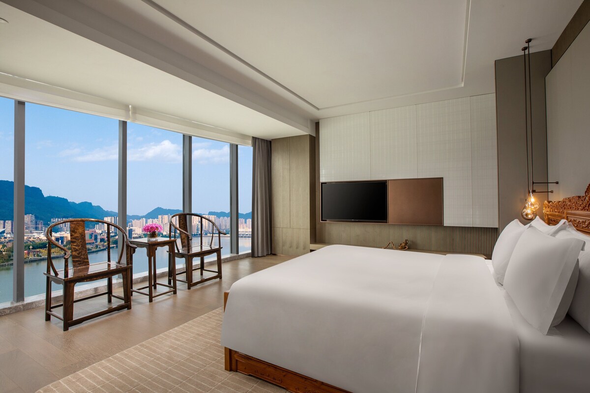 A modern hotel room with a large bed, a TV mounted on the wall, and a view of a cityscape through large windows.
