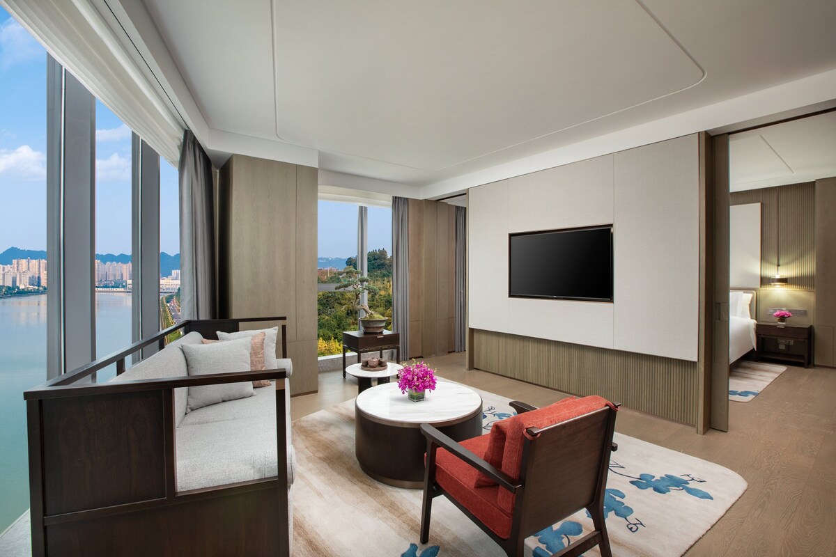 A modern hotel room with a large window offering a city view, a flat-screen TV mounted on the wall, a comfortable seating area with a sofa and armchair, and a small round table with a vase of flowers.