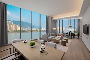 Een moderne woonkamer met een groot raam, een bank, een eettafel met stoelen en een aan de muur bevestigde televisie.