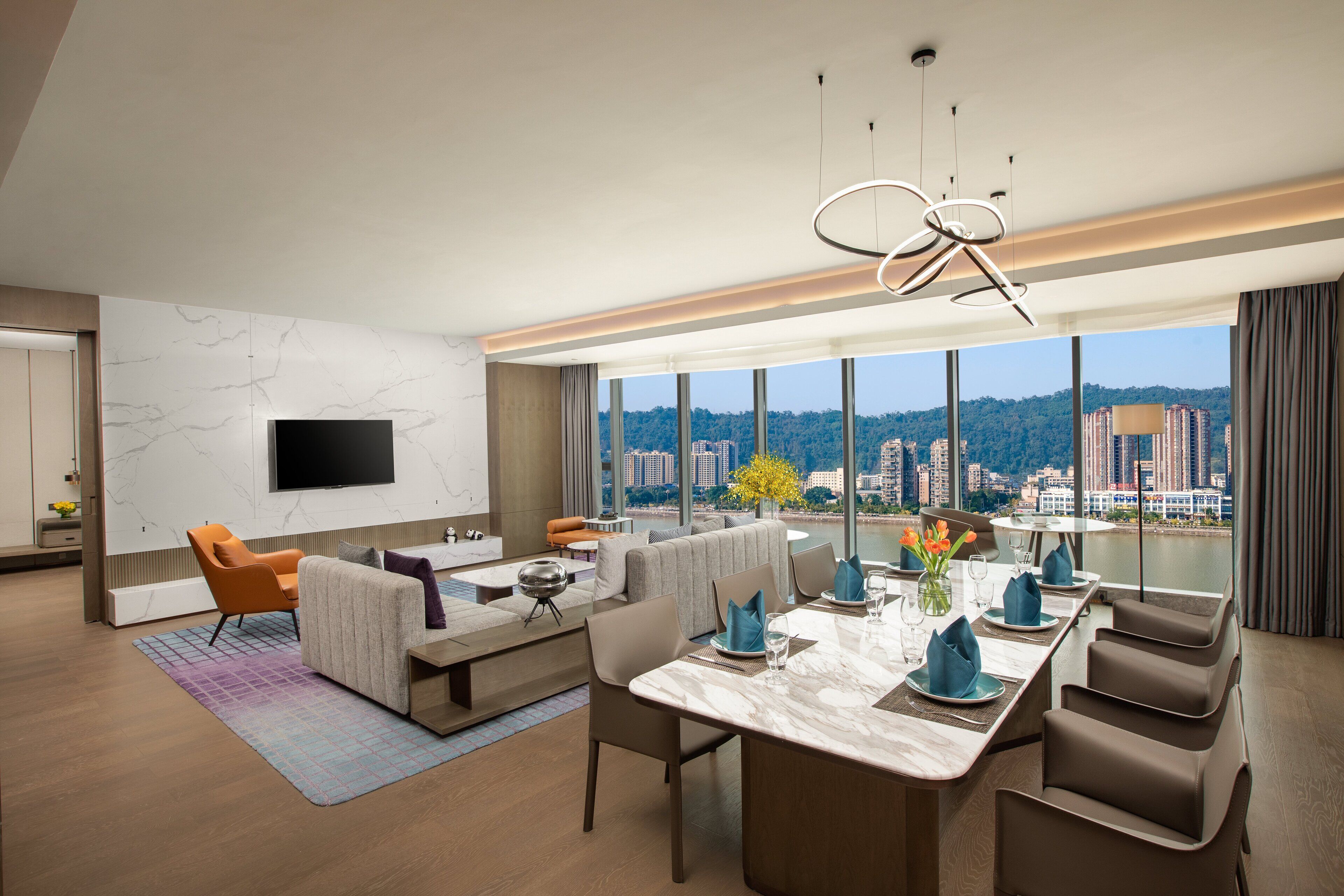 Ein modernes Hotelzimmer mit großem Esstisch für vier Personen, einer Couch, einem an der Wand montierten Fernseher und Blick auf die Stadtlandschaft.
