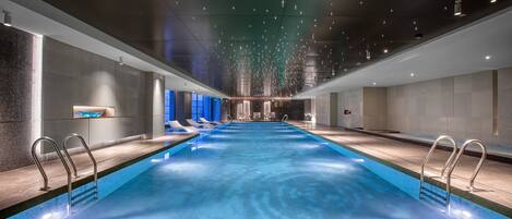 A modern indoor swimming pool with clear water, lounge chairs, and a long bench.