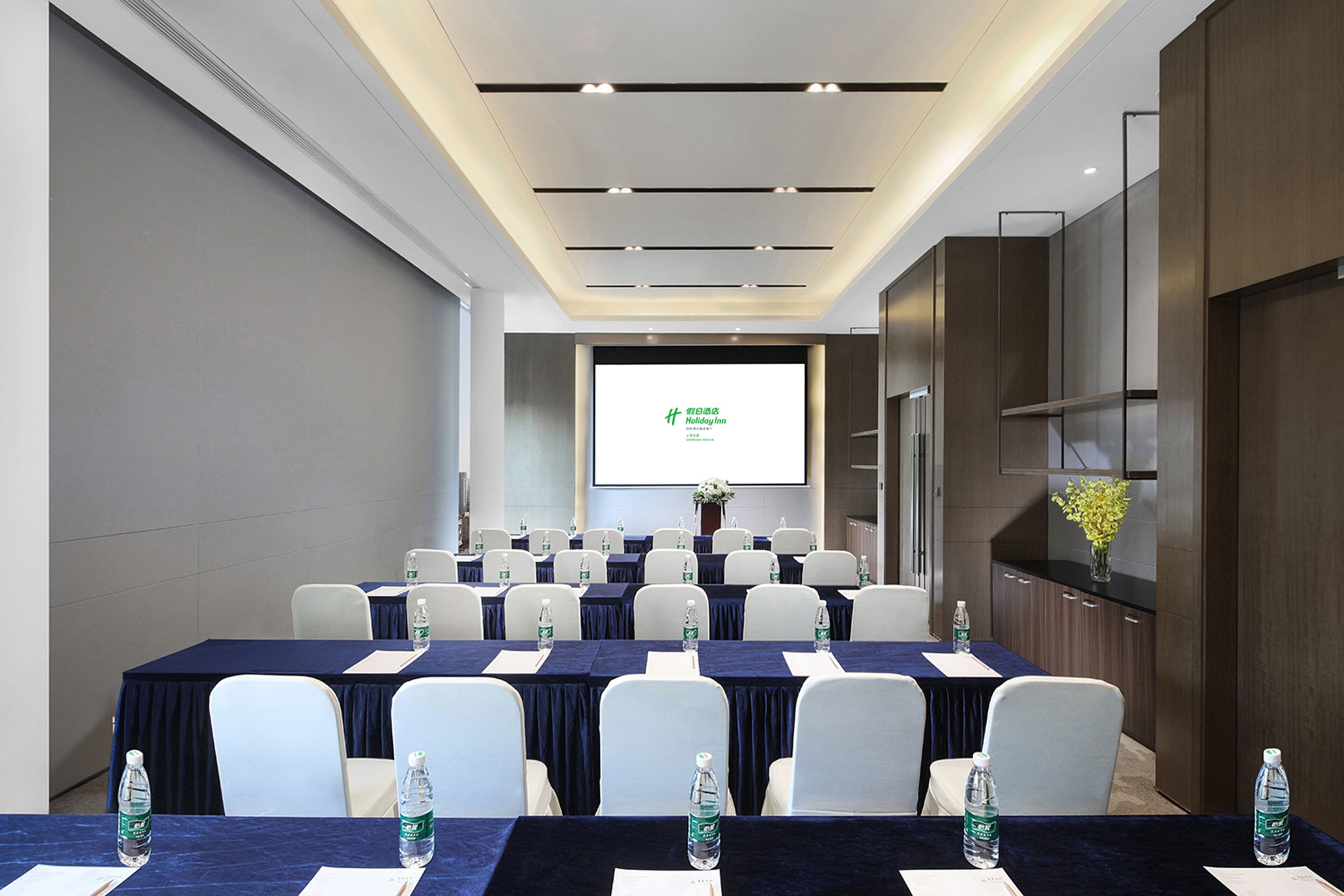 A conference room with a large screen displaying a presentation, rows of tables with chairs, water bottles, and a vase with flowers on the right side.