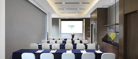 A conference room with a large screen displaying a presentation, rows of tables with chairs, water bottles, and a vase with flowers on the right side.