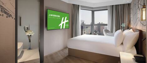 Une chambre d’hôtel avec un grand lit, un téléviseur fixé au mur, une vue sur la ville par la fenêtre et une table de chevet avec une lampe.
