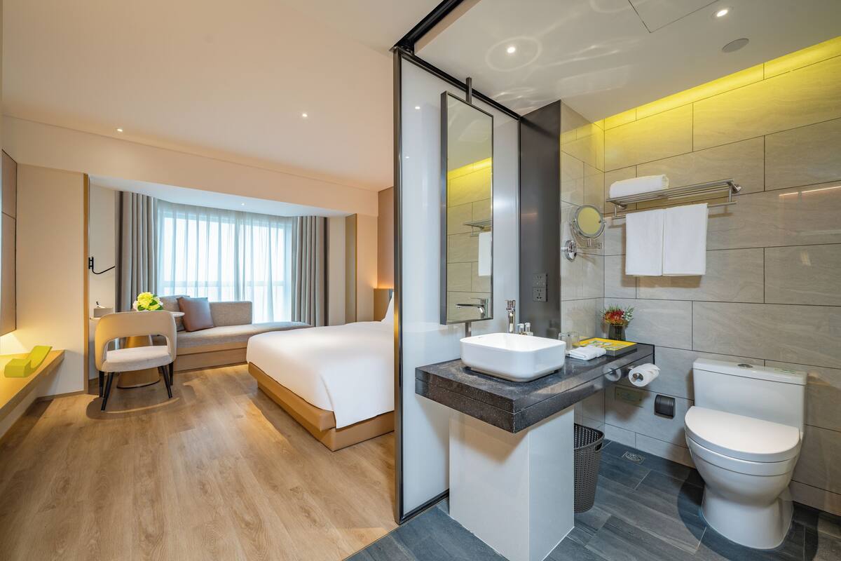 A modern hotel room with a bed, a chair, a bathroom with a sink, mirror, and toilet.
