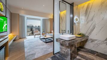 Una habitación de hotel moderna con una cama grande, un baño con tocador de mármol, vistas a la ciudad y un televisor a la izquierda.