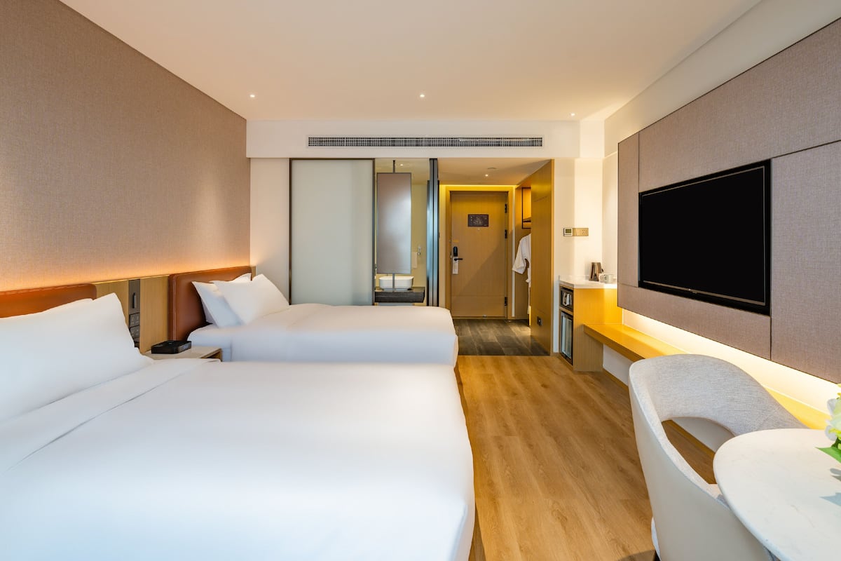 A hotel room with two beds, a TV, a desk with a chair, and a bathroom visible through a door.