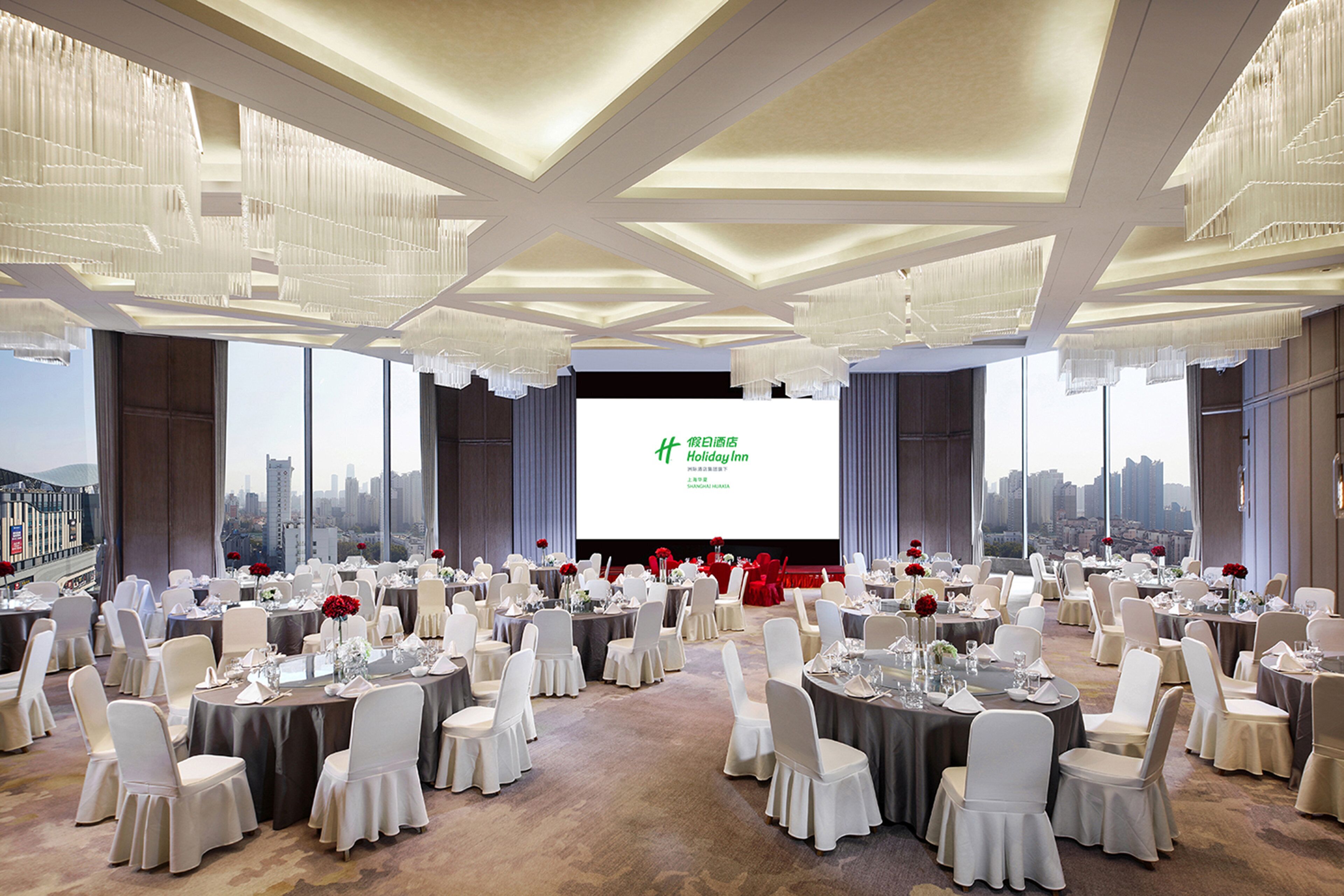 A banquet hall with round tables set for a formal event, large windows offering a city view, and a projection screen displaying text.
