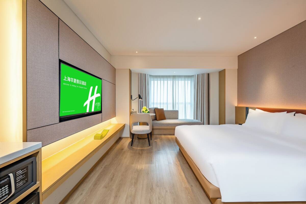 A hotel room with a large bed, a desk with a computer, a TV mounted on the wall, and a sofa.