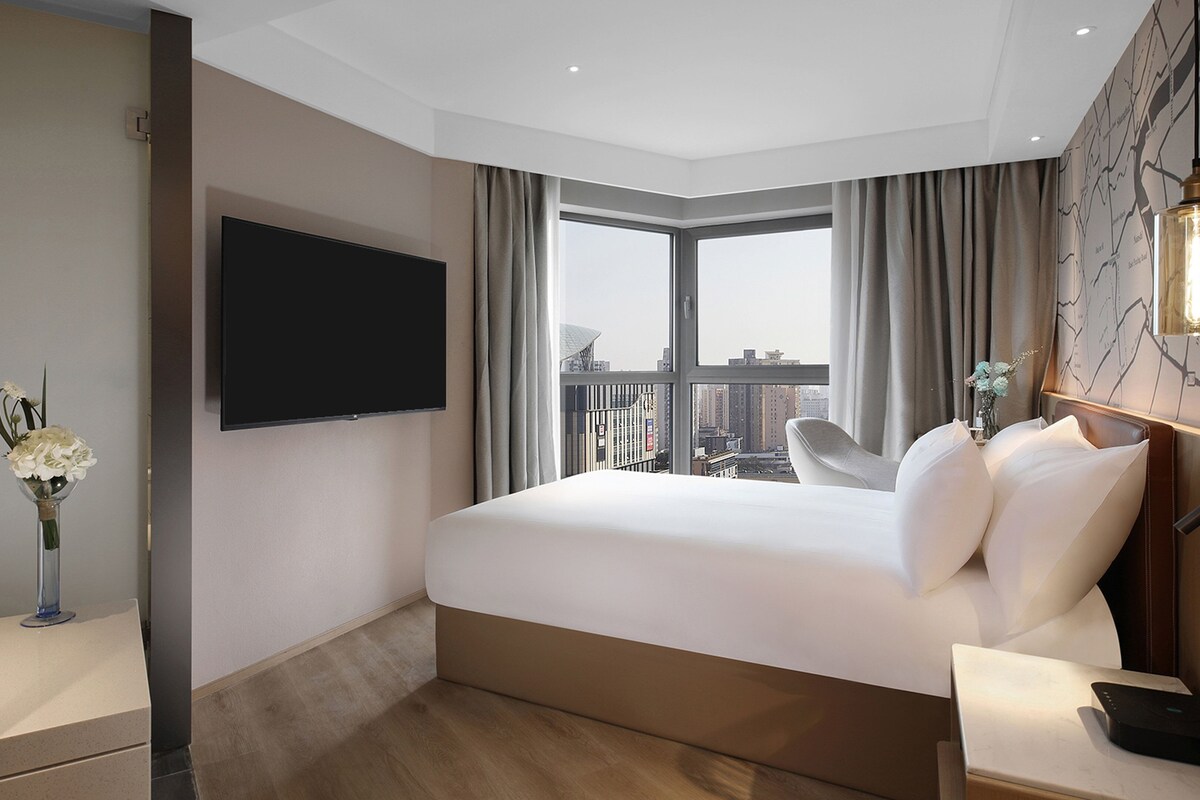 Una habitación de hotel moderna con una cama grande, un televisor de pantalla plana en la pared, vistas a la ciudad a través de un ventanal amplio y una mesita con un florero.