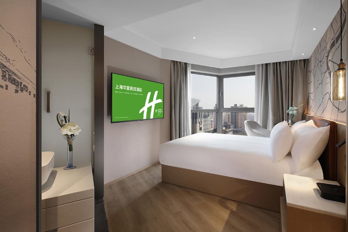 A hotel room with a large bed, a TV mounted on the wall, a view of the city through the window, and a nightstand with a lamp.