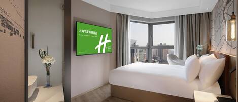 Ein Hotelzimmer mit einem großen Bett, einem an der Wand montierten Fernseher, Blick auf die Stadt durch das Fenster und einem Nachttisch mit Lampe.