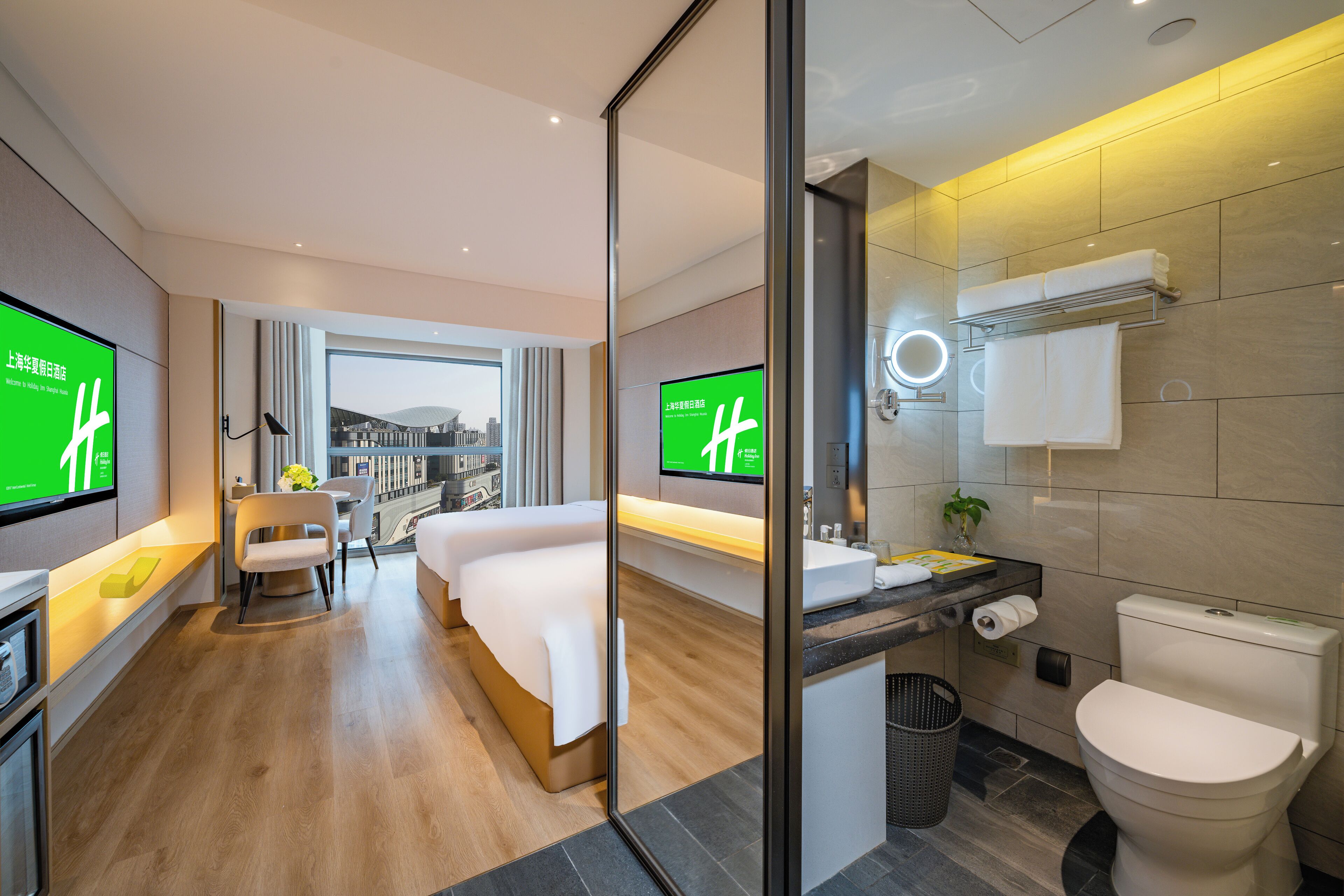 A modern hotel room with a bed, a desk, a TV, a bathroom with a toilet, and a view of the city.