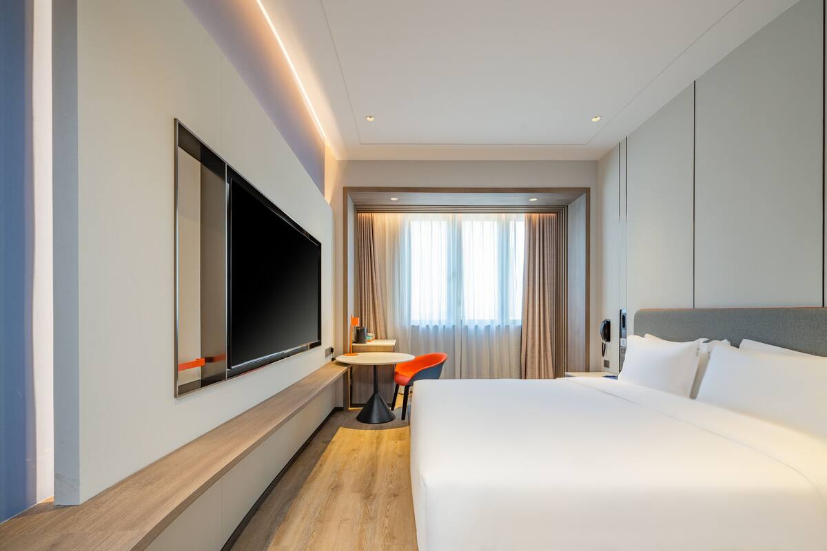 A modern hotel room with a large bed, a flat-screen TV mounted on the wall, a small round table with a chair, and a window with curtains.