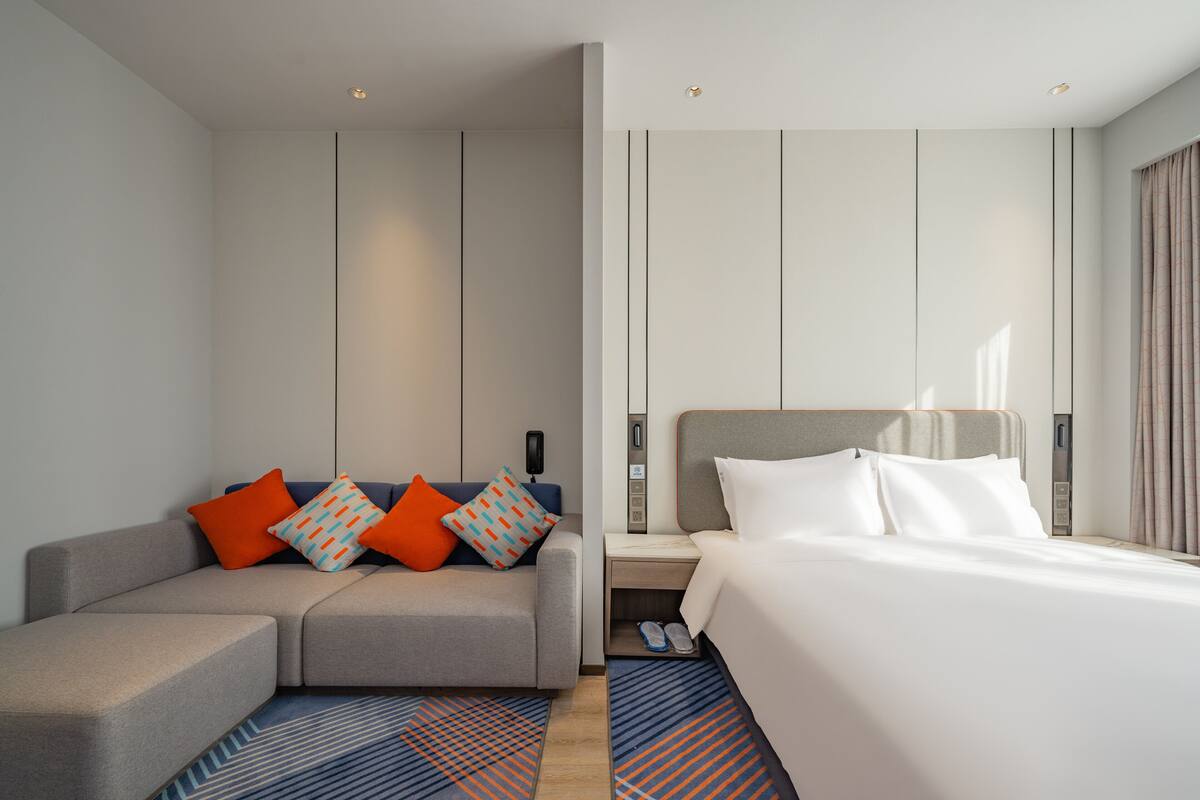 A modern hotel room with a grey sofa, a bed with white linens, and a nightstand with a lamp.