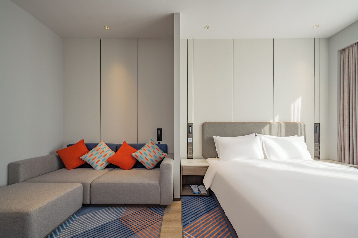 A modern hotel room with a grey sofa, a bed with white linens, and a nightstand with a lamp.