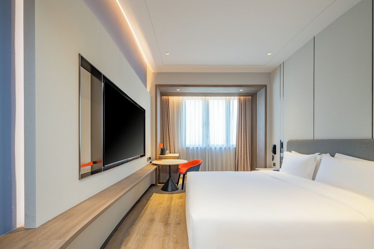 A modern hotel room with a large bed, a flat-screen TV mounted on the wall, a small round table with a chair, and a window with curtains.