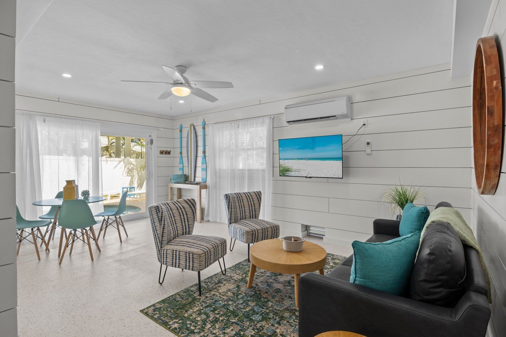 A modern living room with a ceiling fan, a flat-screen TV, and a view of a beach through the window.