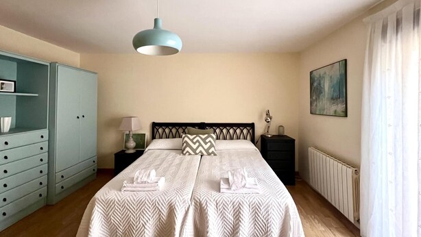Un dormitorio con cama, armario, mesita de noche, lámpara, un cuadro y ventana con cortinas.