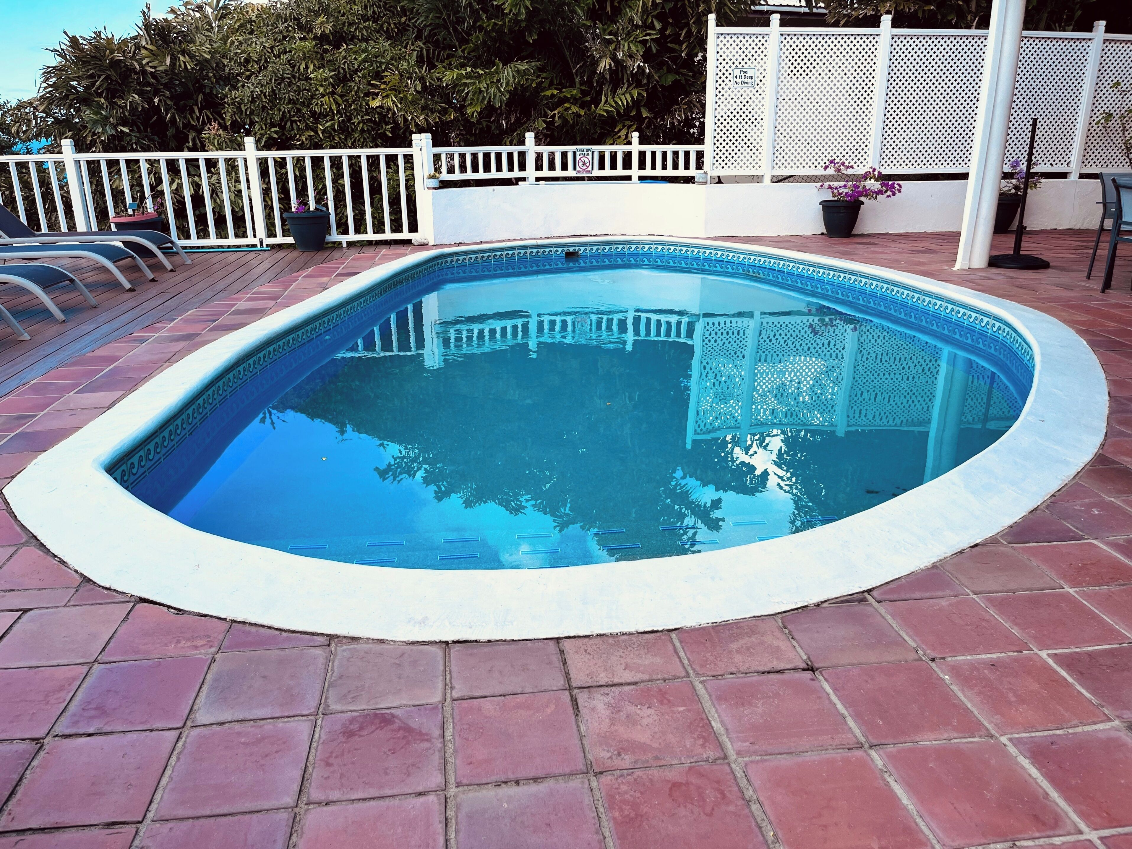 A swimming pool with a white deck, surrounded by a white fence, and potted plants.