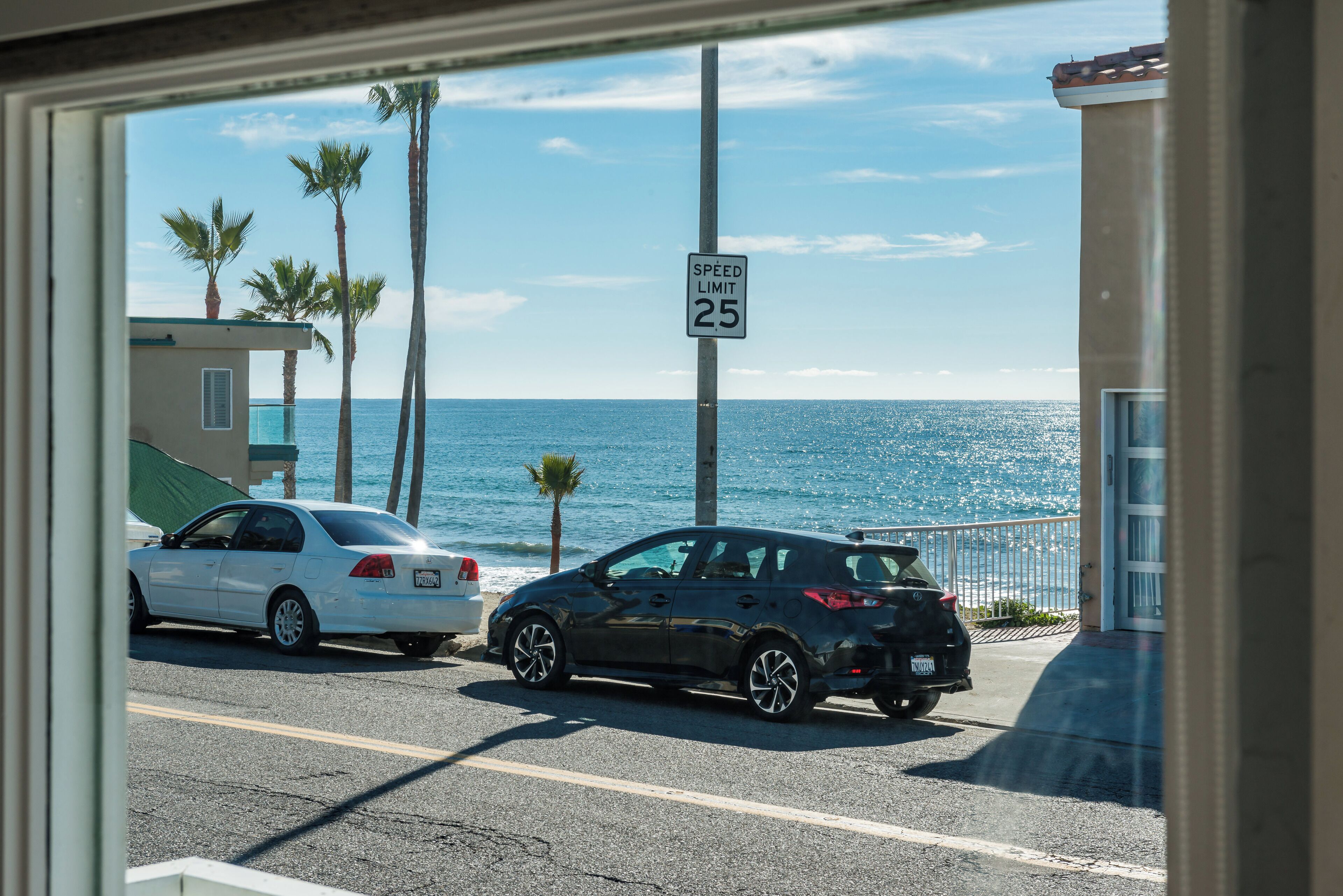 A street view with a speed limit sign of 25, two parked cars, palm trees, and a view of the ocean.