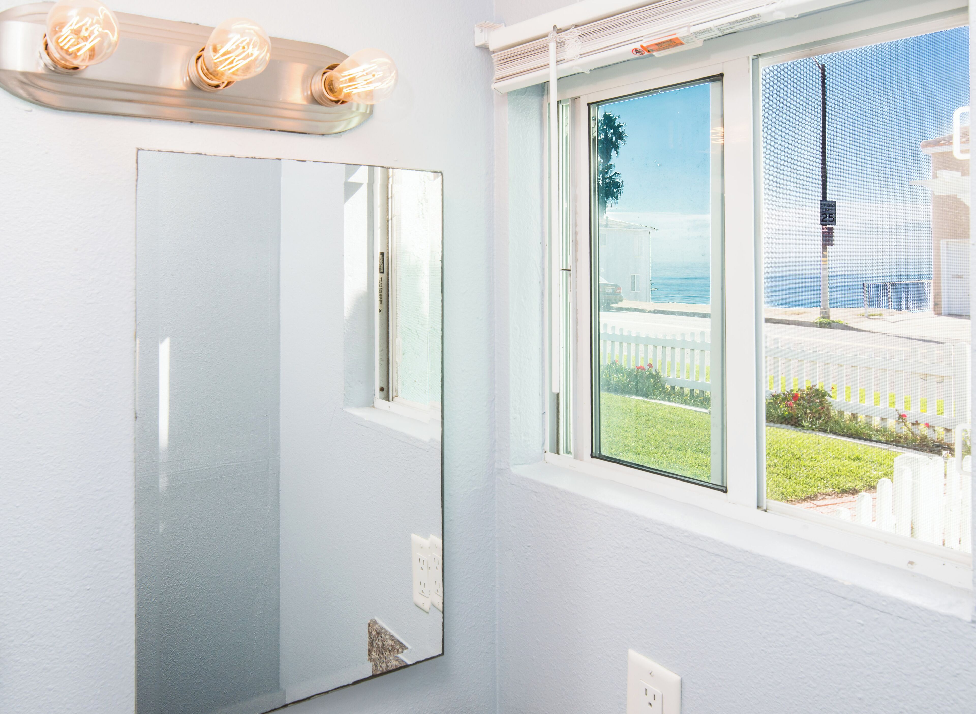 A room with a large mirror, a window with a view of the ocean, and a light fixture with three bulbs.