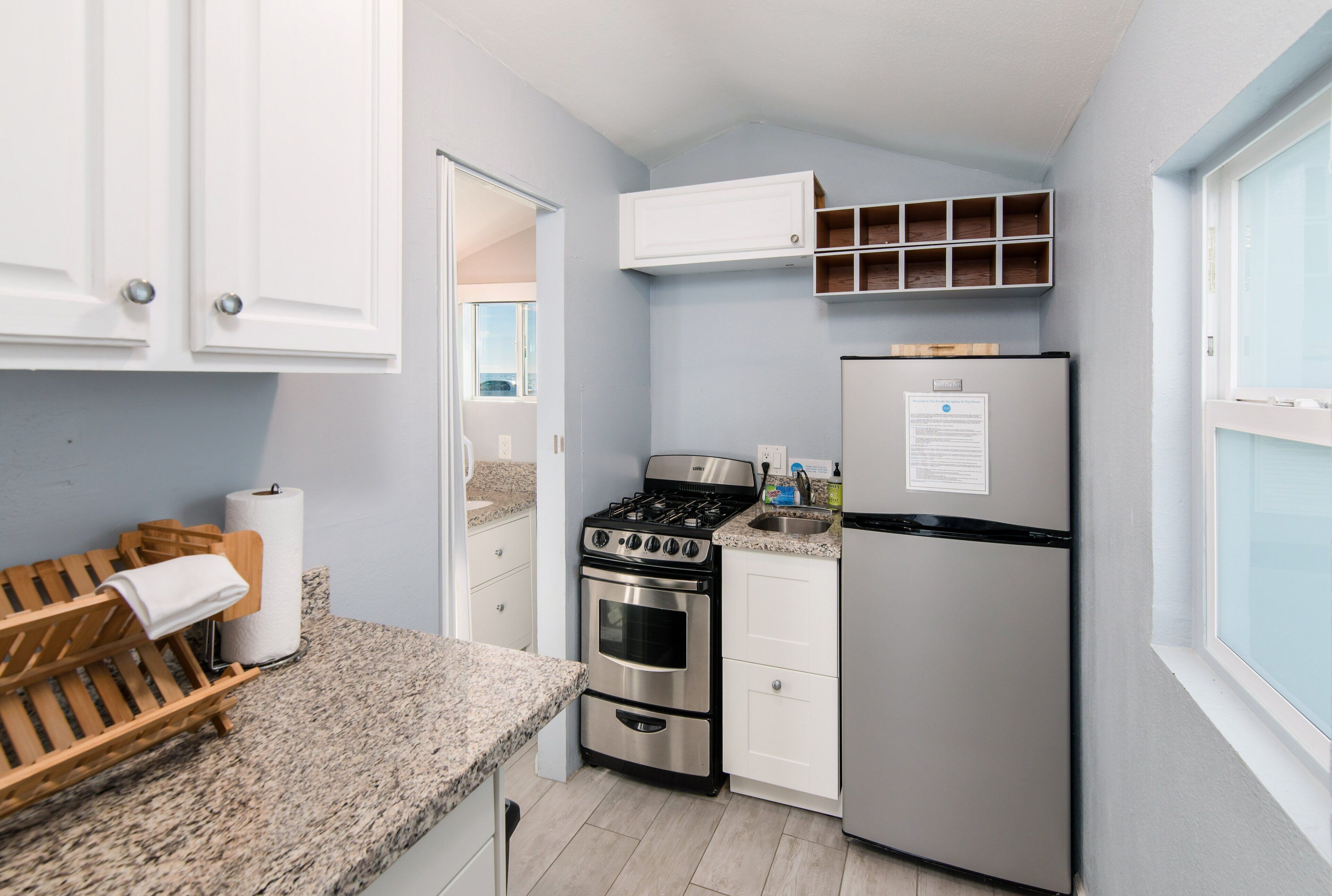 A compact kitchen with a gas stove, oven, and refrigerator, wooden bench, and white cabinets.