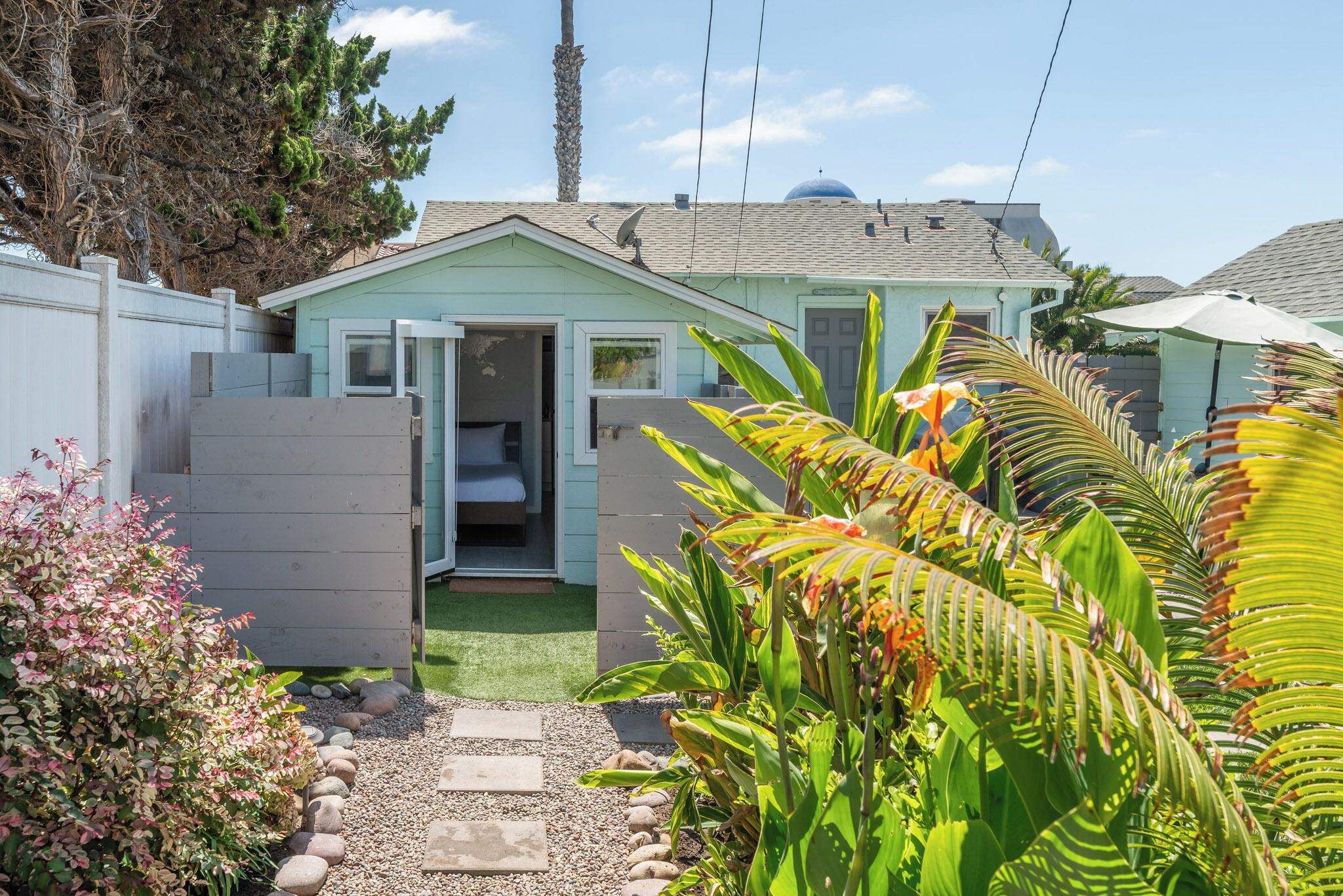 A small, light green house with an open door, a gravel path, and tropical plants.