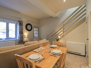 Un área de comedor con una mesa de madera dispuesta para cuatro personas, un sofá, un reloj y una escalera.