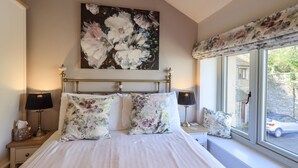 A bedroom with a large bed, floral bedding, a painting above the bed, and a window with a view of the outside. - Cringol Cottage (Ambleside)