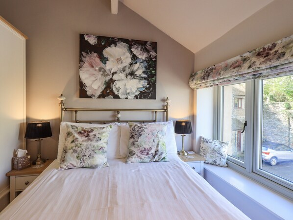 A bedroom with a large bed, floral bedding, a painting above the bed, and a window with a view of the outside.